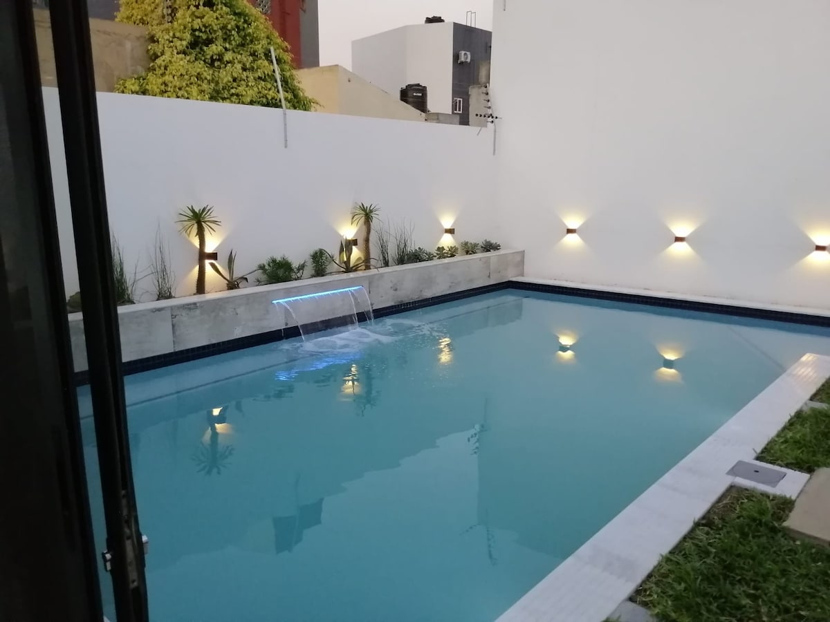A tranquil private pool is showcased, featuring softly illuminated water and a gentle waterfall feature. Surrounding greenery complements the sleek, minimalistic design. Warm lighting on the walls highlights the serene atmosphere of the outdoor space.