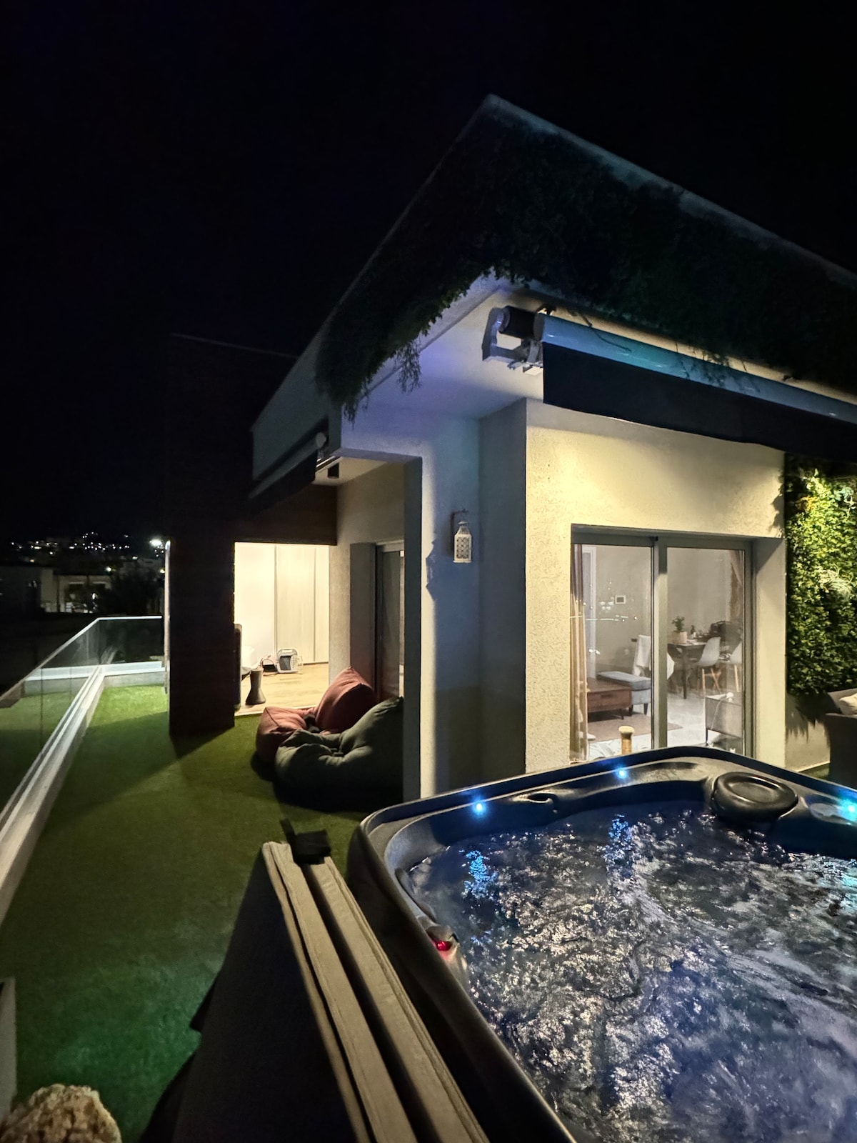 A tranquil outdoor space is illuminated by soft lighting, featuring a jacuzzi and areas for relaxation. A green wall adds a natural touch alongside the sleek architecture. The inviting ambiance creates a perfect setting for evening enjoyment.