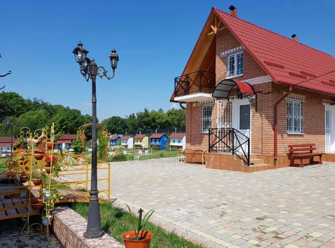 A brick house with a red roof and decorative balcony sits in a well-maintained outdoor area. Paved pathways and vibrant green foliage create a welcoming environment. Street lamps and flower pots enhance the space, while additional colorful buildings are visible in the background.
