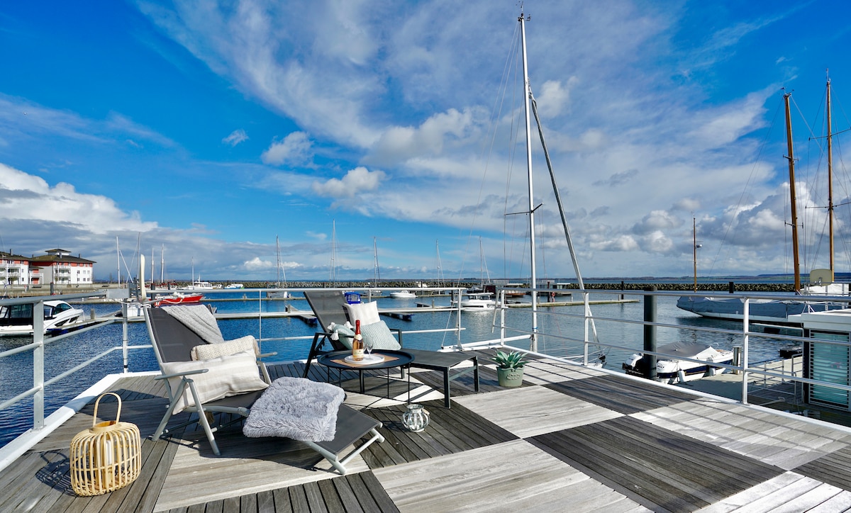 An outdoor deck offers a serene view of the water, showcasing boats docked nearby. Lounge chairs are positioned alongside a small table, with a cozy blanket and a plant accenting the space. The sky is bright with scattered clouds, enhancing the waterfront ambiance.