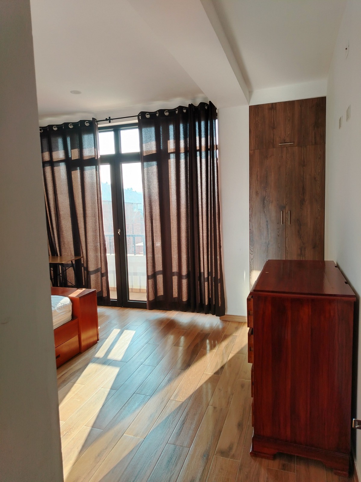 A well-lit bedroom is shown, featuring large windows dressed with dark curtains. Light streams in, illuminating the wooden flooring. A dresser stands against the wall, while a bed is visible in the corner of the room. The overall space feels inviting and organized.