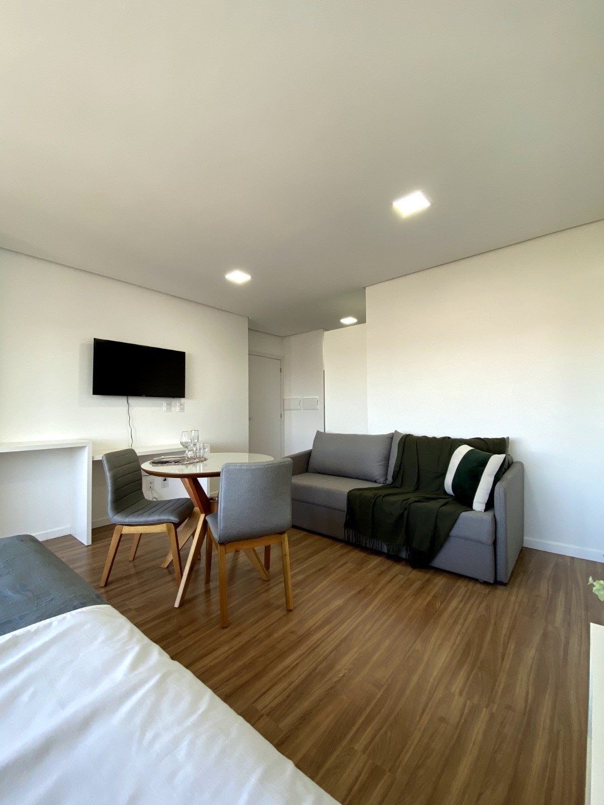 A modern studio space features a cozy gray sofa and a minimalist dining table set with four chairs. A wall-mounted television is positioned for easy viewing, and natural light enhances the warm wooden floor. A throw blanket adds a touch of comfort.