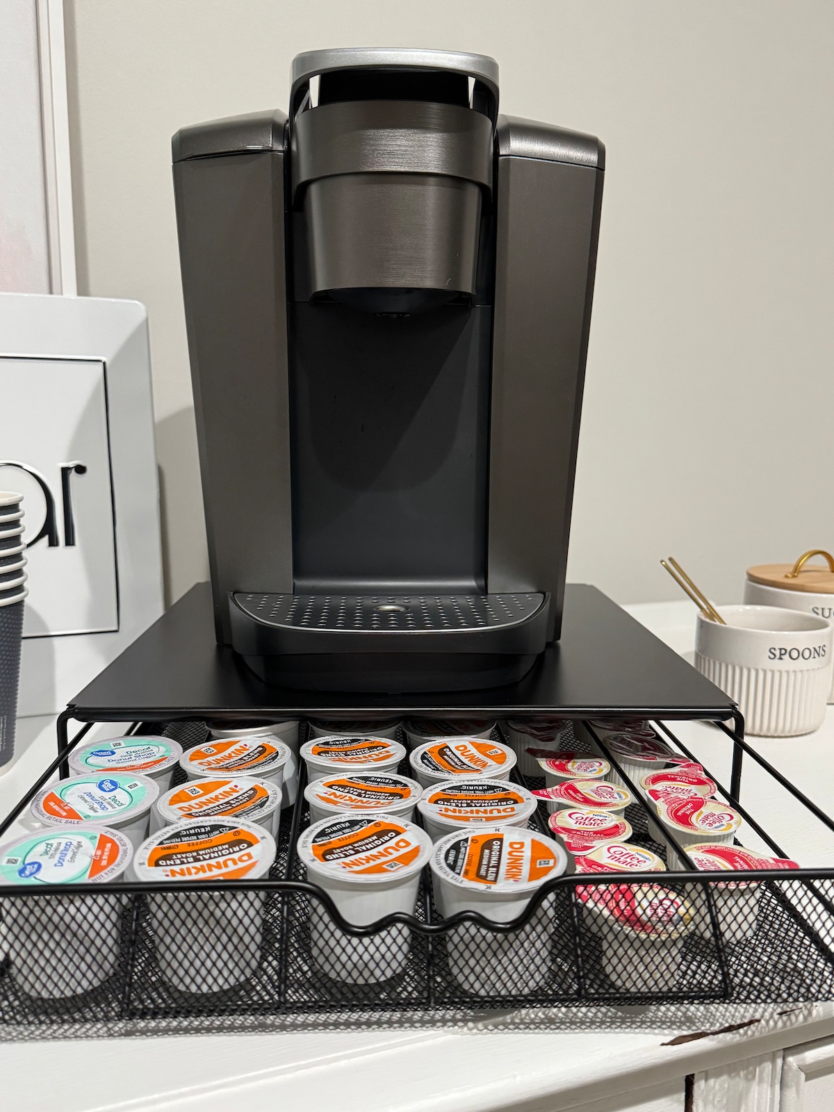 A modern Keurig coffee maker is positioned on a black shelf, surrounded by an organized assortment of coffee pods. The pods are neatly arranged in a wire basket below, showcasing various flavors and creamers for guest convenience.