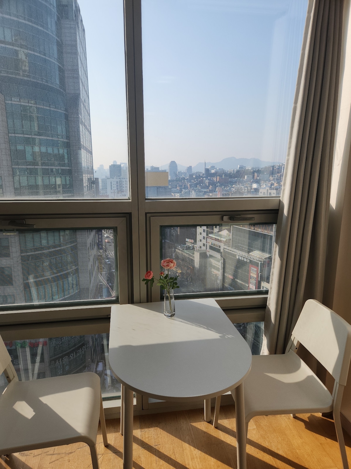 A small dining area is located by a large window, offering a view of the city skyline. A round table is accompanied by two white chairs, and a vase with fresh flowers is placed at the center, enhancing the inviting atmosphere.