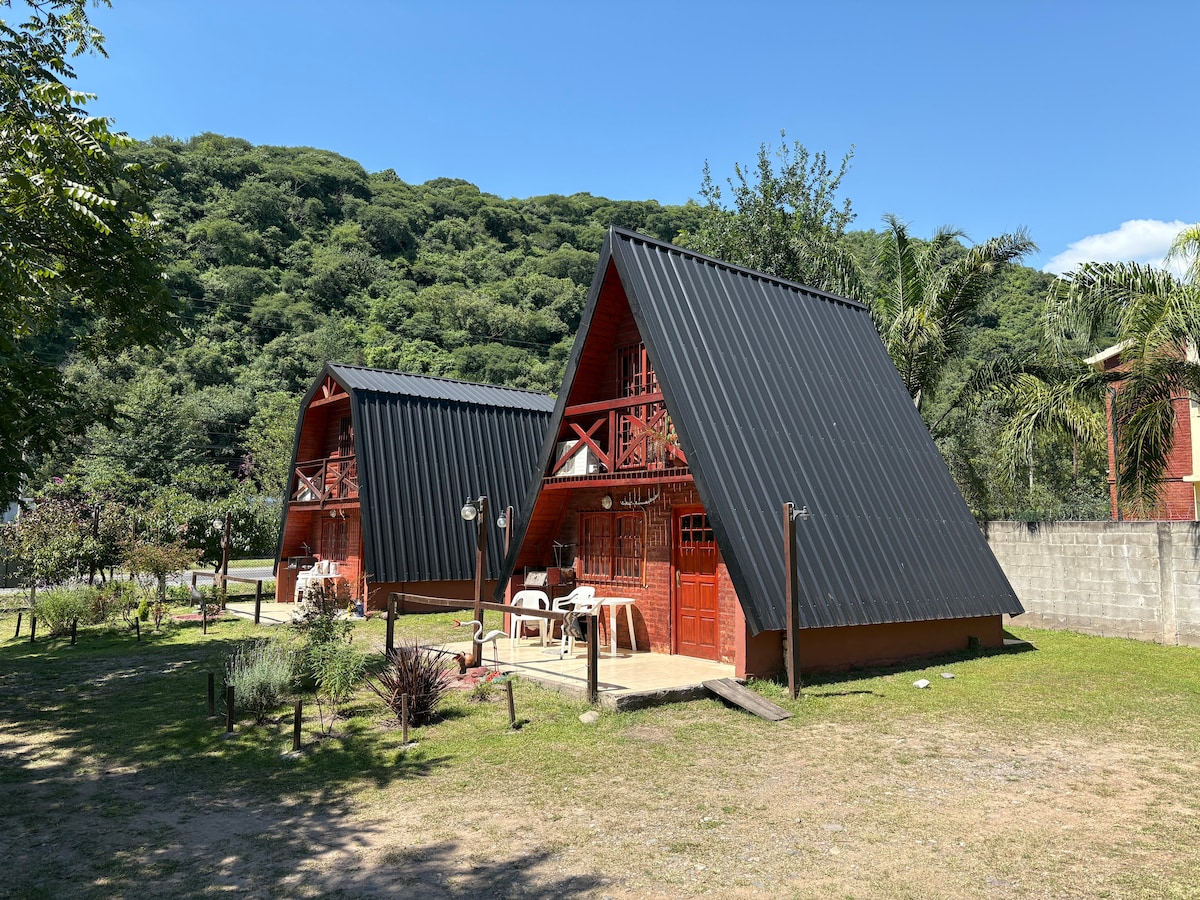 Two independent cabins with striking triangular roofs are located amidst lush greenery. Each cabin features a welcoming porch area with seating arrangements. The scenic landscape includes gentle hills and plants, creating a serene environment for relaxation.