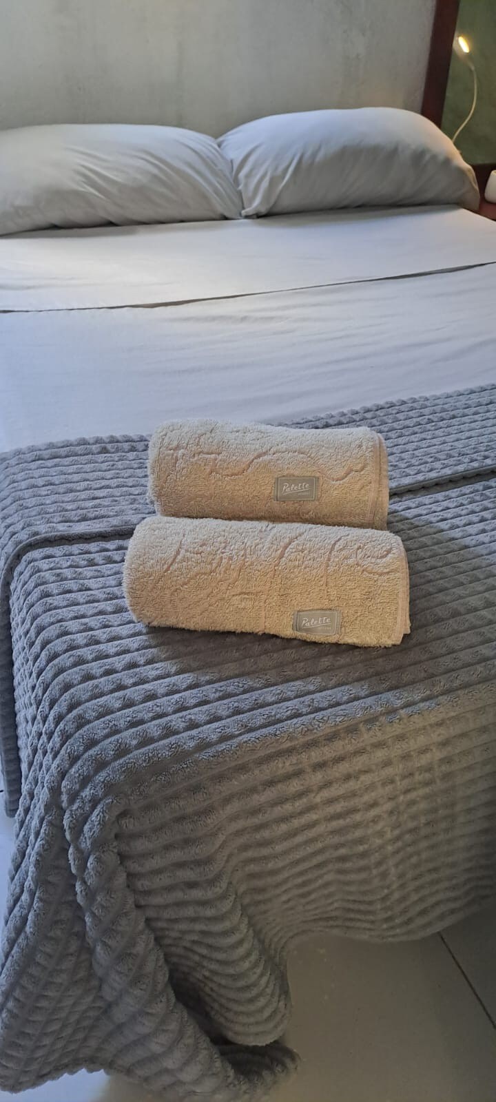 A neatly arranged bed is presented with a soft, neutral-toned blanket. Two rolled towels are placed on the bedding, showcasing a textured design. The room features a clean and minimalist aesthetic, with ample natural light contributing to the serene atmosphere.