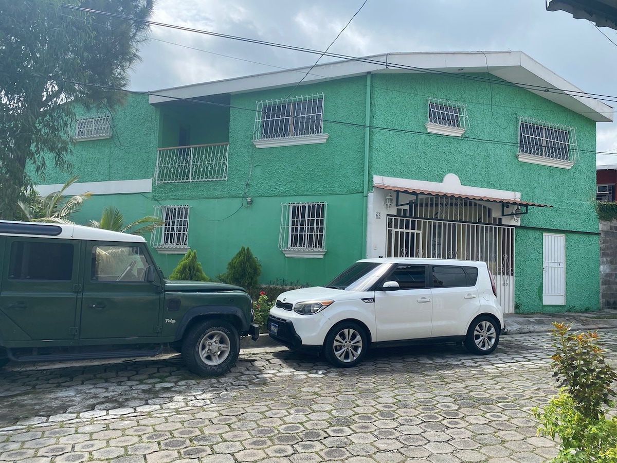 A two-story green house features a distinctive exterior with white accents. Windows decorated with metal grilles allow natural light inside. Two vehicles are parked in front, with cobblestone paving leading to the entrance. Surrounding greenery enhances the overall setting.