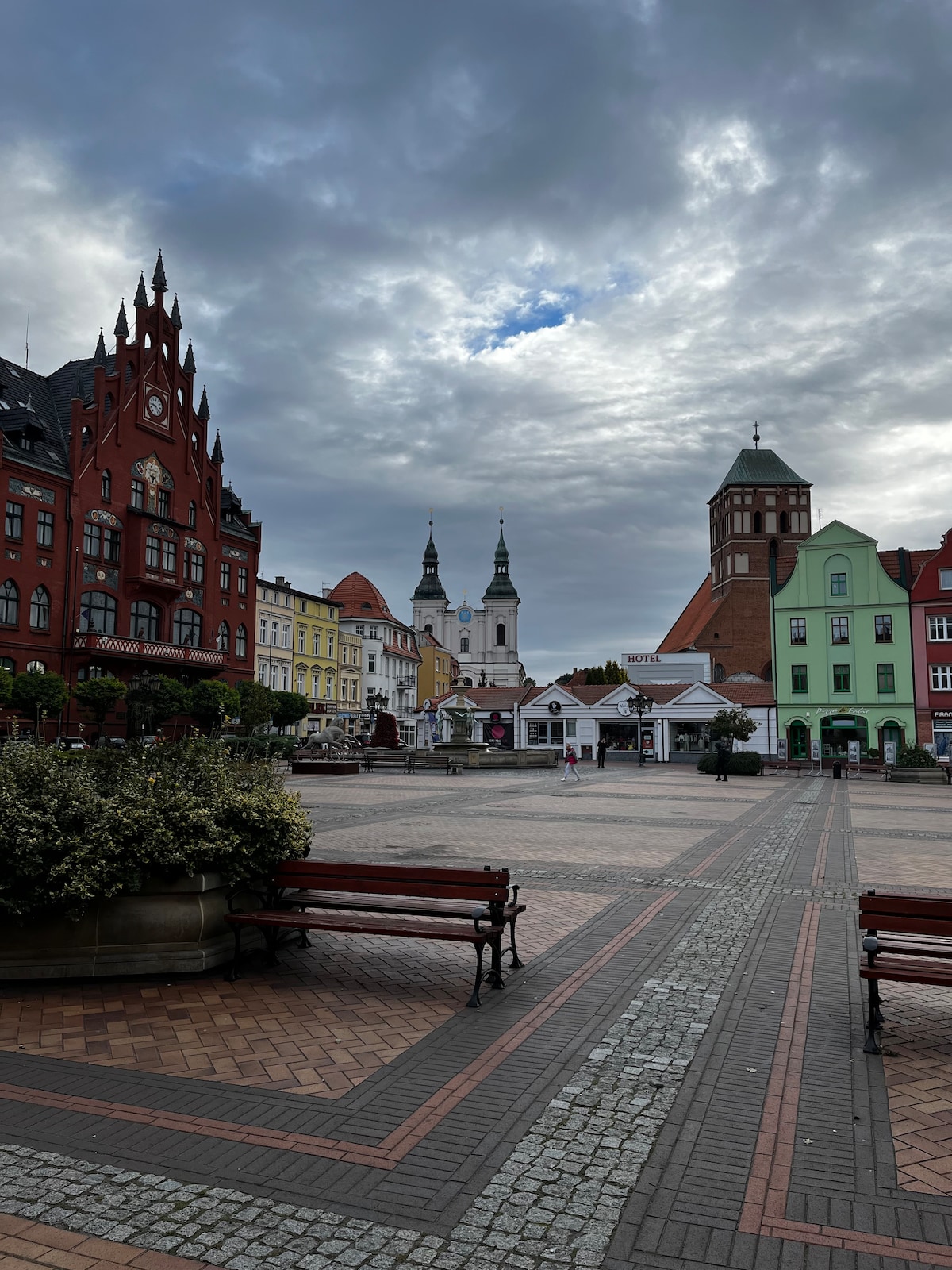 A vibrant town square is framed by colorful historic buildings, including a red Gothic-style structure and a green facade. Benches line the cobblestone pathway, offering a place to sit. The sky is overcast, adding depth to the scene while the spires of nearby churches rise in the background.