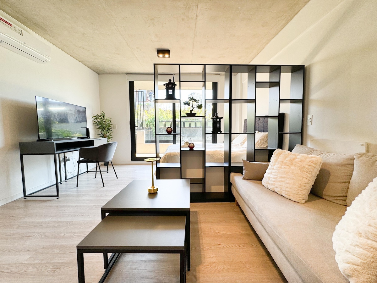 A modern living space showcases an open layout with a cozy sofa positioned near a sleek coffee table. A decorative room divider adds privacy, while a desk and television are visible. Sunlight enters through large glass doors leading to an outdoor area.