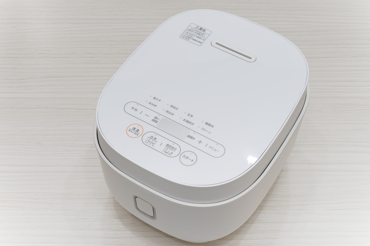 A sleek white rice cooker is displayed on a light wood surface. The device features a control panel on the top with various buttons and settings clearly marked. Its modern design is complemented by a compact shape, making it a functional addition to the kitchen.