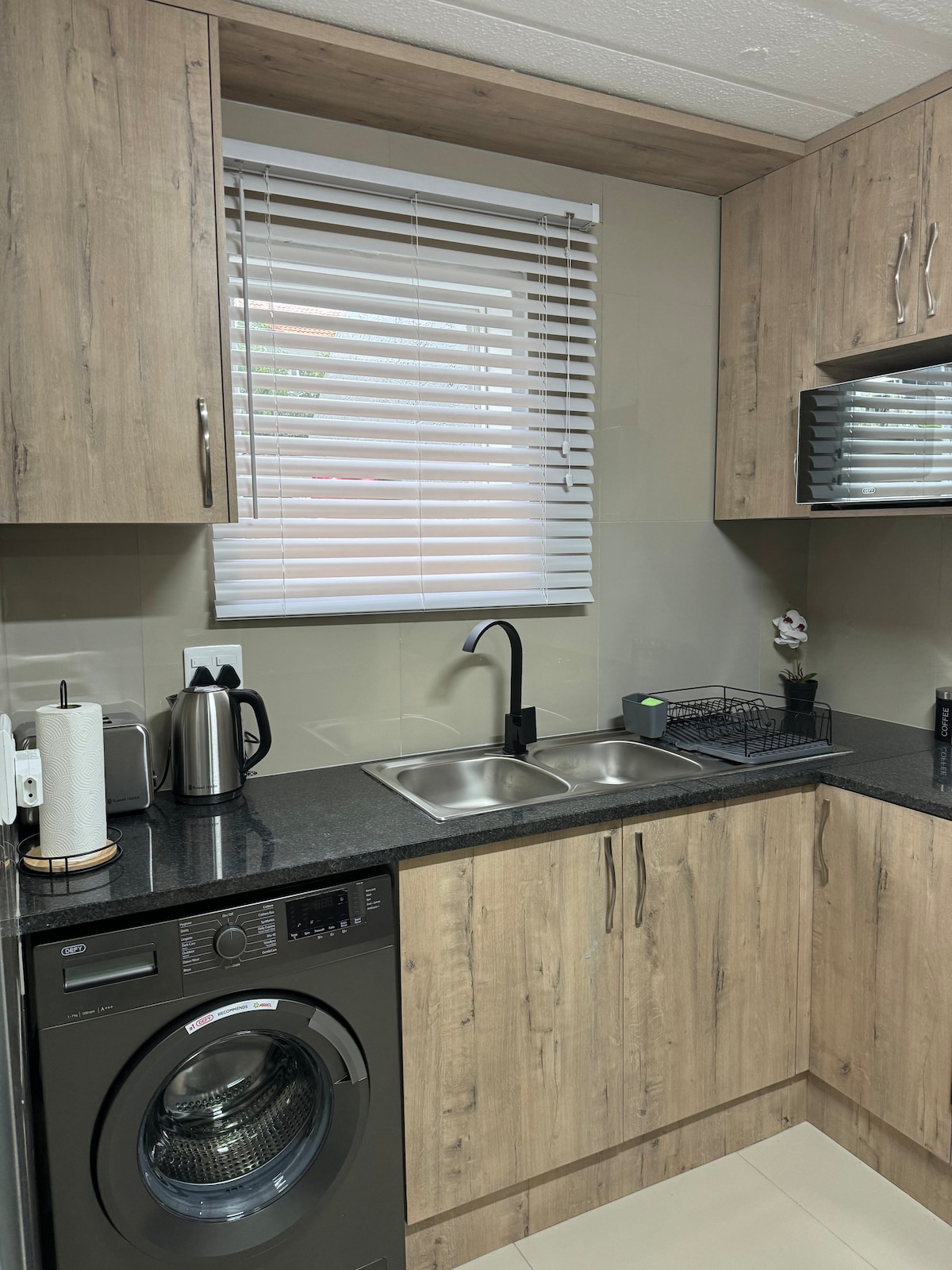 A modern kitchen features light wooden cabinetry and dark countertops. A sleek black faucet is positioned over a double sink, accompanied by essential kitchen appliances including a kettle and a washing machine. Natural light filters through a window with white blinds.