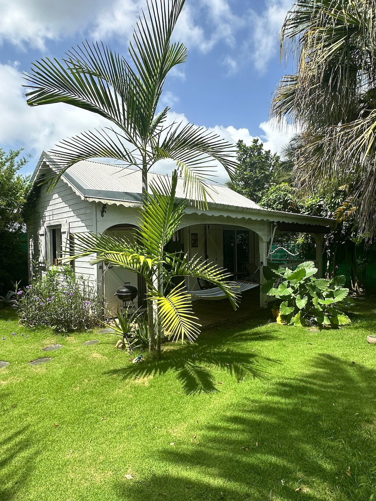 A traditional Creole house is framed by lush greenery and tropical plants. A small porch is visible, providing shaded seating. The building features a gabled roof and a light-colored exterior, blending harmoniously with the vibrant garden surrounding it.