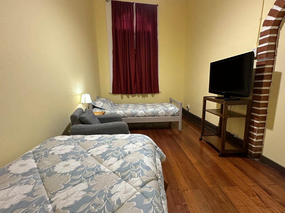 A room featuring two beds, one of which is positioned with a gray seating area nearby. A television stands on a wooden table against the wall. Natural light enters through a window, partially obscured by burgundy curtains. The floor is made of polished wood.