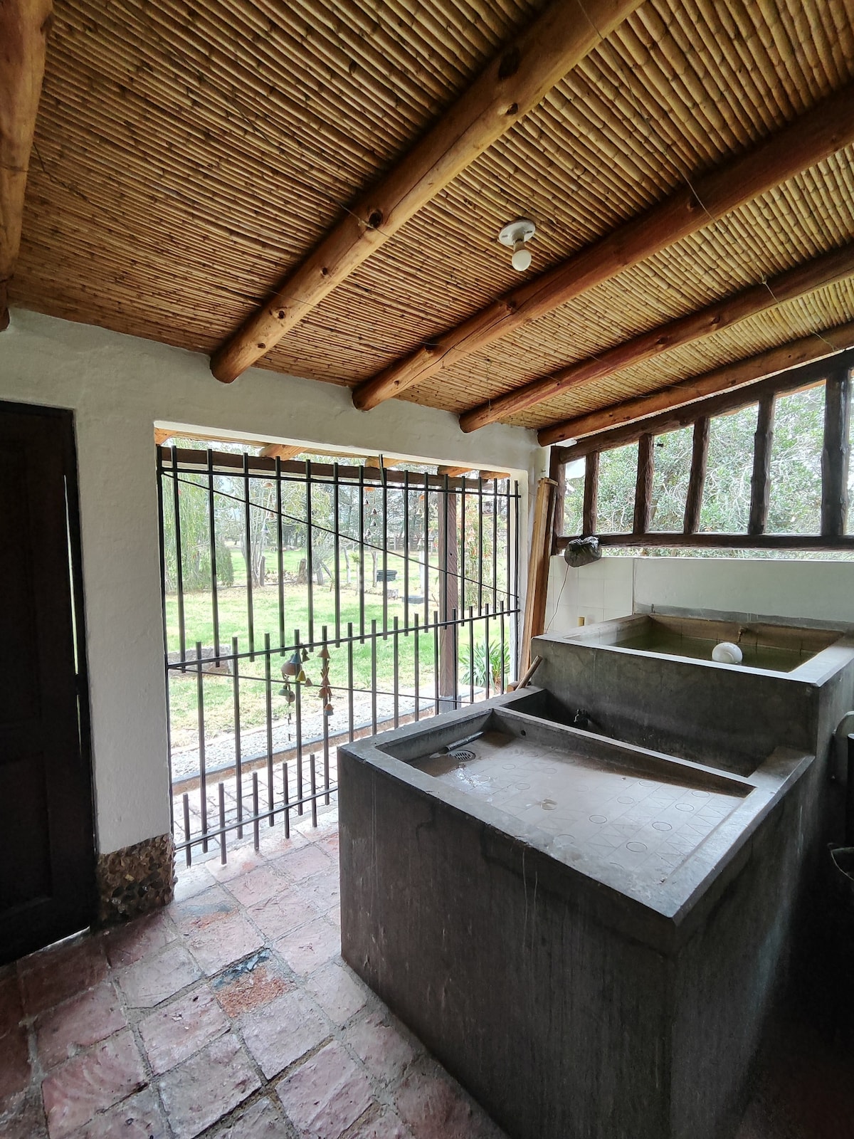 A utility area is equipped with a stone sink and a spacious countertop. Natural light enters through multiple windows, complementing the rustic wooden beams overhead. A barred window provides a view of the surrounding greenery, enhancing the sense of openness in the area.