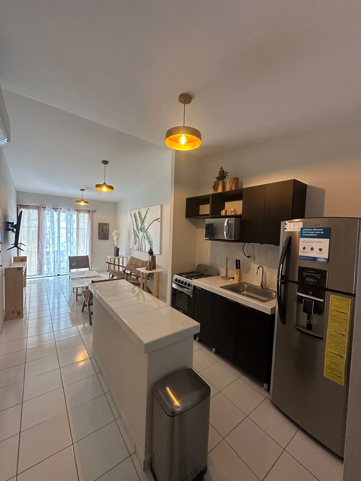 A modern kitchen is depicted, featuring sleek black cabinetry and stainless steel appliances. A central island provides additional counter space. Natural light filters through large windows dressed with light curtains, enhancing the open layout. Dining area includes wooden chairs and a table, creating a welcoming space.
