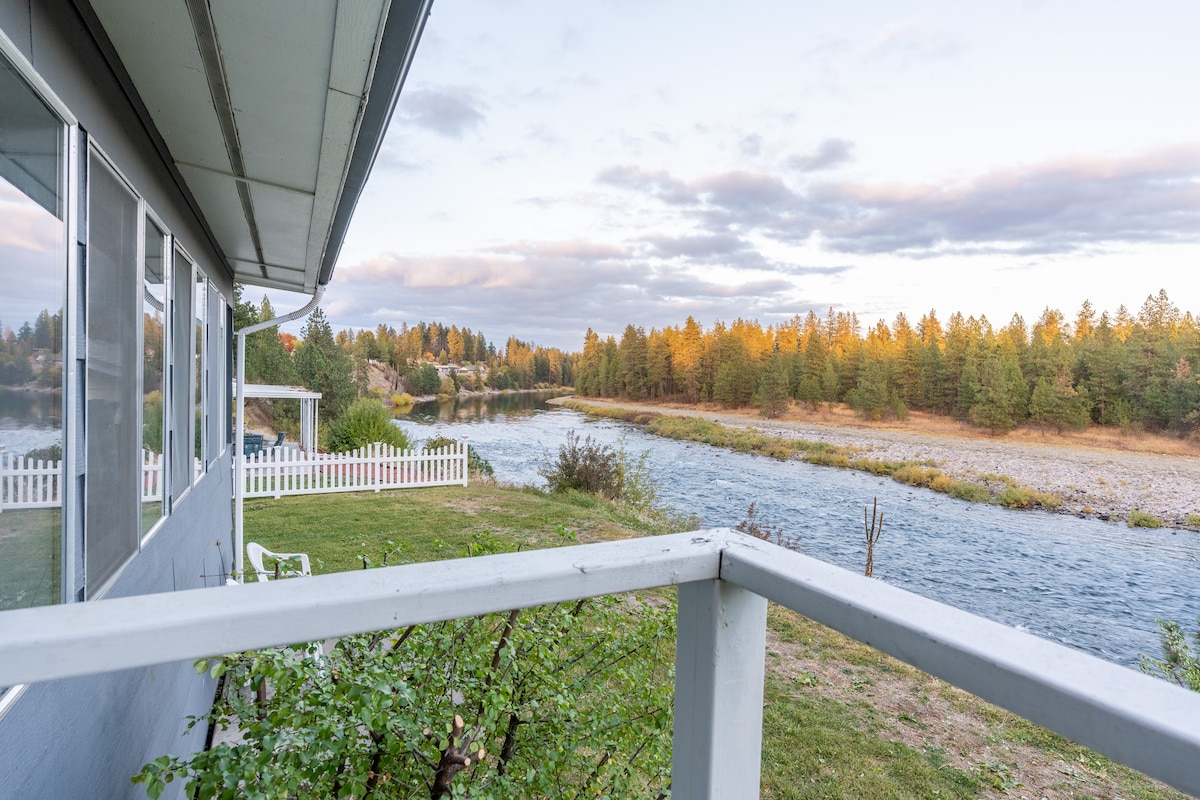A scenic view of the Spokane River is framed by greenery and trees. The image showcases a tranquil riverbank with a gentle current, while a white fence borders the yard. Soft hues of sunset illuminate the landscape, enhancing the natural beauty of the surroundings.