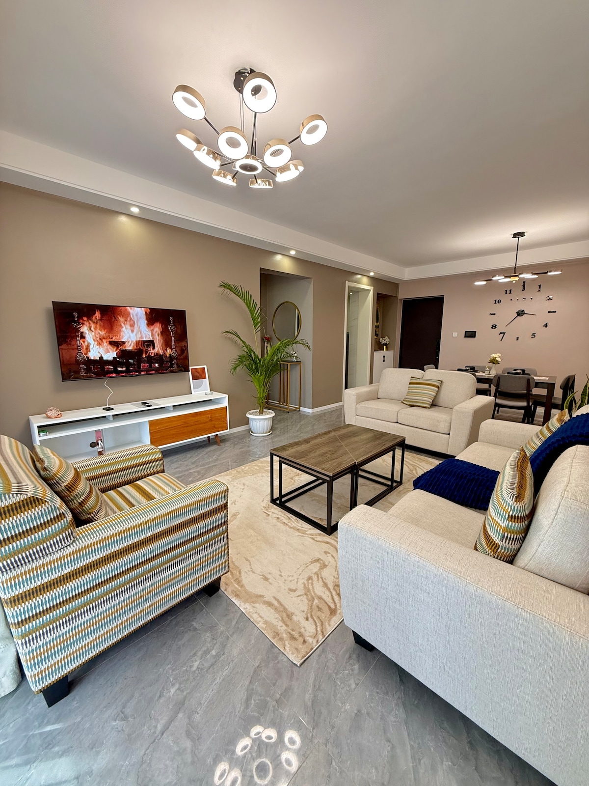 A spacious living area is showcased, featuring comfortable seating options, including two striped chairs and a neutral sofa. A coffee table sits centrally, complemented by a flat-screen TV displaying a fireplace scene. Soft lighting and elegant decor enhance the modern design.