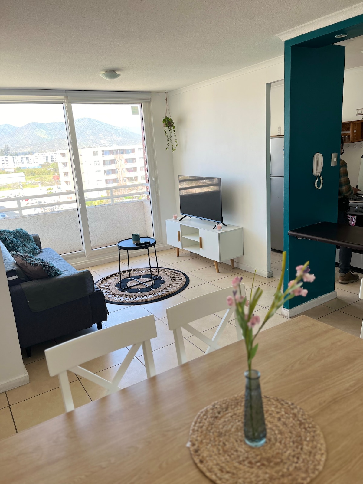A bright living area is visible, featuring a television on a low white stand and a comfortable dark sofa. A round table with four white chairs occupies the dining space. A small plant adorns the table, and large windows offer views of distant mountains.