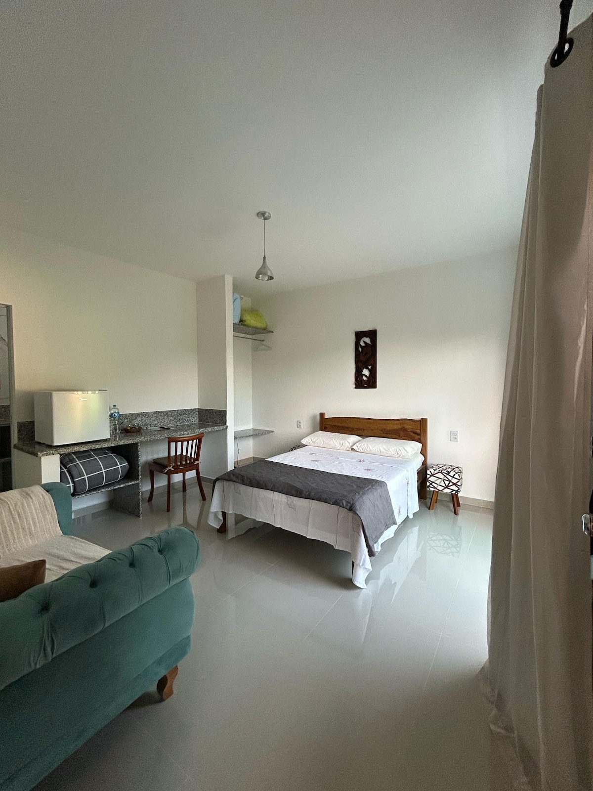 A well-lit room features a spacious bed with white linens and a dark throw blanket. A small table and chair are positioned nearby, alongside a kitchenette equipped with a countertop and a miniature fridge. A cozy green sofa adds comfort to the space.