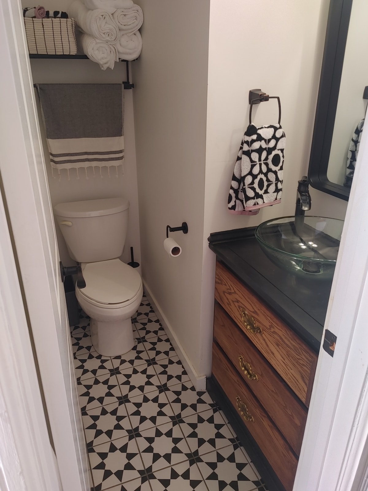 A compact bathroom features a white toilet and a dark surface vanity with a glass sink. Decorative towels are neatly arranged on a rack. A black and white patterned tile floor adds visual interest, while a mirror is mounted above the sink for convenience.