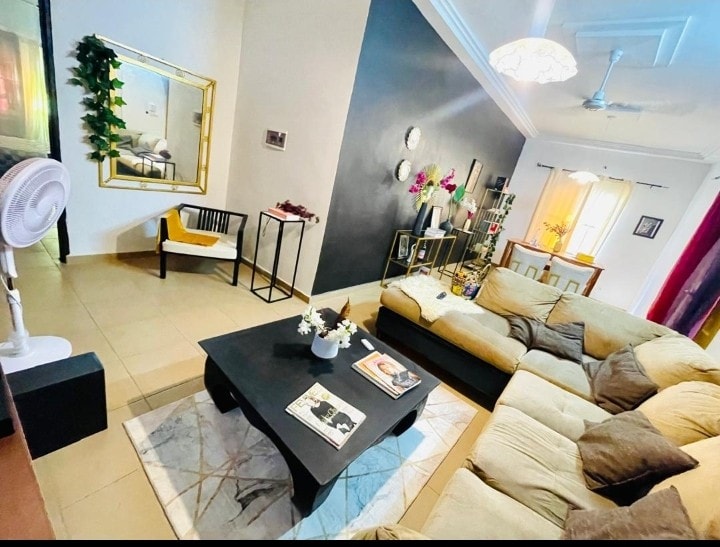 A spacious living area is depicted featuring a large, L-shaped sectional sofa and a coffee table. A mirror with a golden frame is mounted on the wall, while decorative plants add vibrancy. Natural light enters through the window, highlighting the room's modern decor.