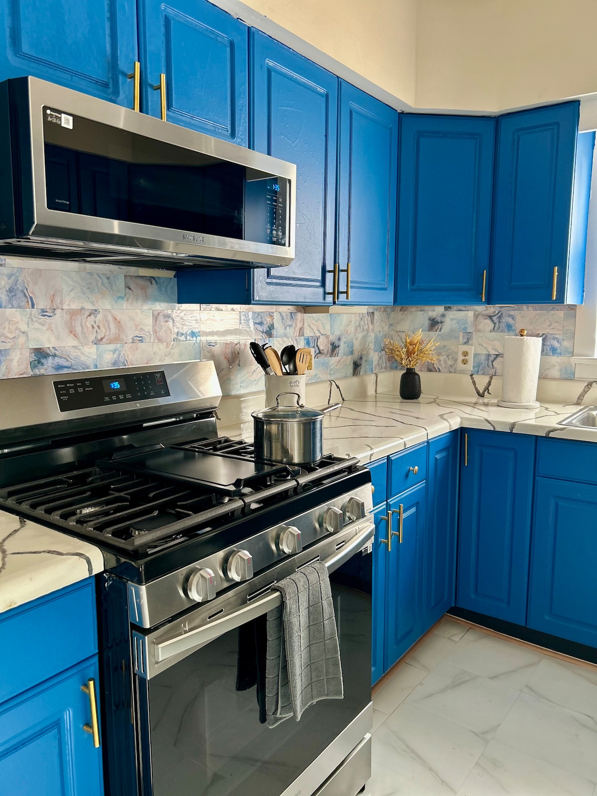 A modern kitchen features vibrant blue cabinetry complemented by elegant marble countertops. Stainless steel appliances, including a microwave and stove, are neatly arranged. Kitchen utensils and a small pot sit on the stove, while a window allows natural light to brighten the space.