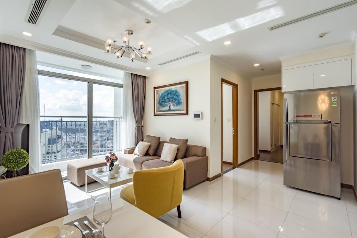 A modern living area is seen with a comfortable sofa and a yellow accent chair, complemented by a large window offering a view of the city. Bright natural light fills the space, highlighting a sleek dining table and a fully equipped kitchen with stainless steel appliances.