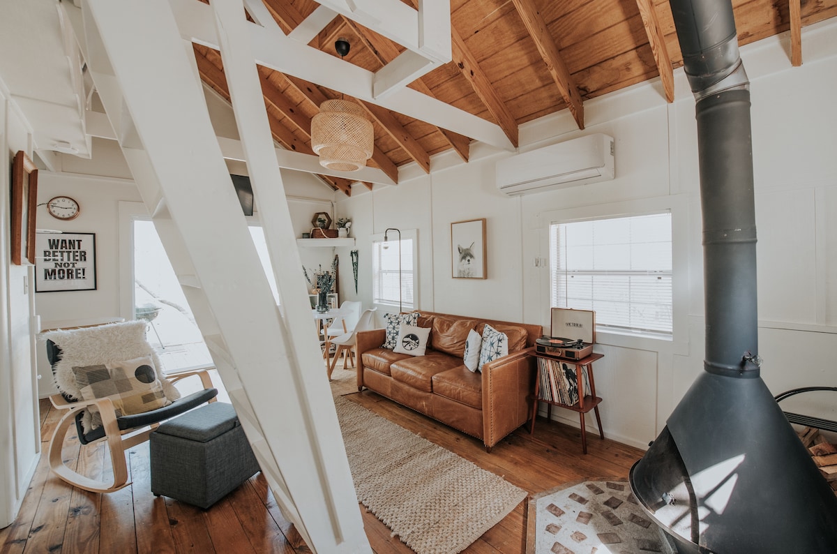 The open living area features exposed wooden beams with natural light streaming through large windows. A leather couch and a cozy lounge chair are situated on warm wood floors, complemented by a woven area rug. A modern stove stands in the corner, adding to the cabin's rustic charm.