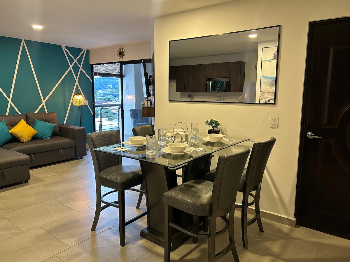 A dining area is positioned next to a spacious living room. A modern glass table is surrounded by four upholstered chairs. A large mirror reflects light and the adjacent kitchen area. Bright accent pillows add color to the nearby sofa, while a balcony is visible through sliding glass doors.