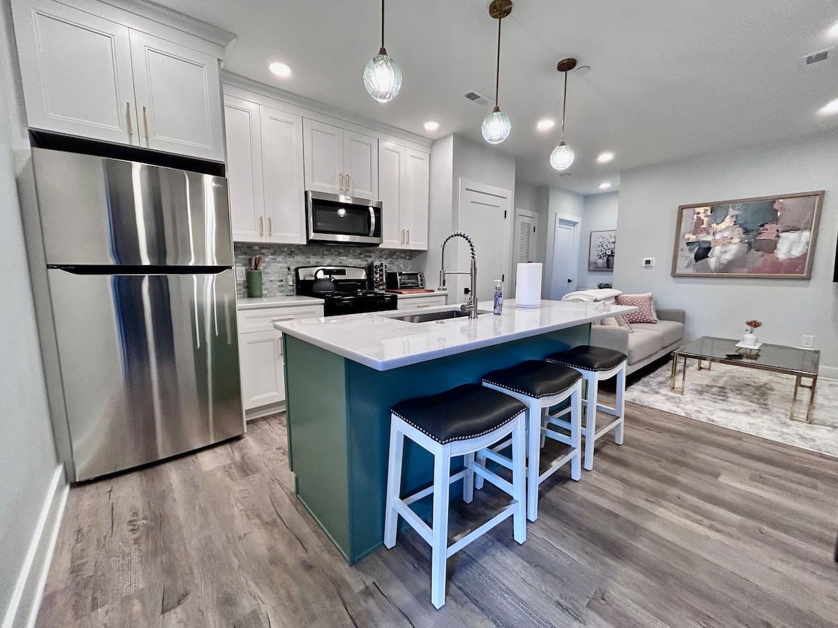 A modern kitchen space is highlighted, featuring stainless steel appliances and a large countertop with jade accents. Three white bar stools are positioned at the island. Soft lighting fixtures suspend above the area, with a glimpse of a cozy seating arrangement in the background.