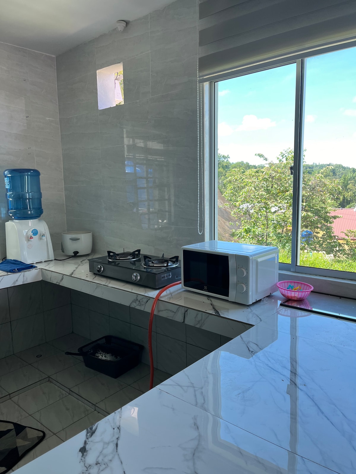 An open concept kitchen features a gas stove and a microwave, positioned on a marble countertop. A water dispenser sits beside a window that allows natural light to illuminate the space. Views of greenery are visible through the window, enhancing the kitchen's inviting environment.