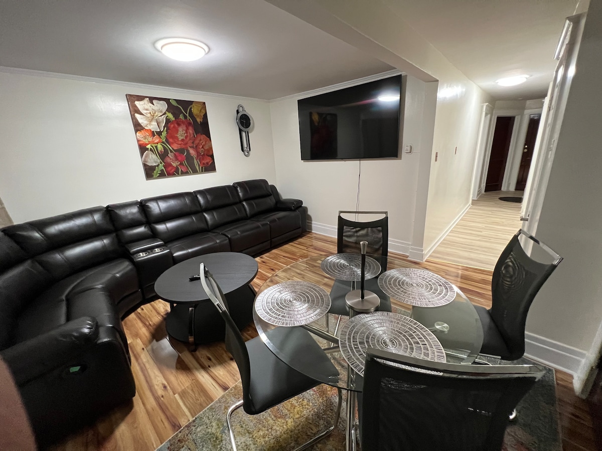 A modern living area features a large sectional sofa facing a wall-mounted television. A round dining table with four chairs is positioned in front of a decorative rug, while floral artwork adds a touch of color to the room.