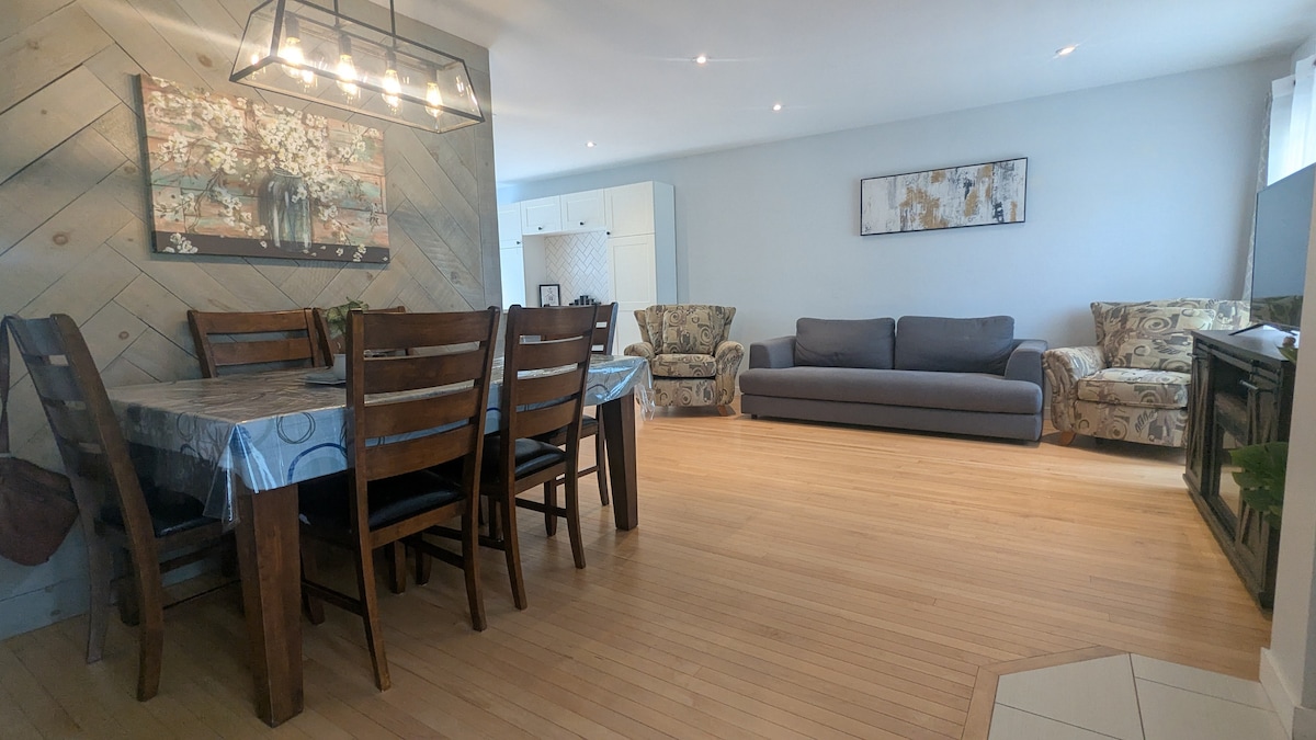 A spacious living area features a dining table with eight wooden chairs and a comfortable gray couch. Two patterned armchairs complement the seating arrangement. Natural light illuminates the bamboo flooring, while light-colored walls provide a calming backdrop.