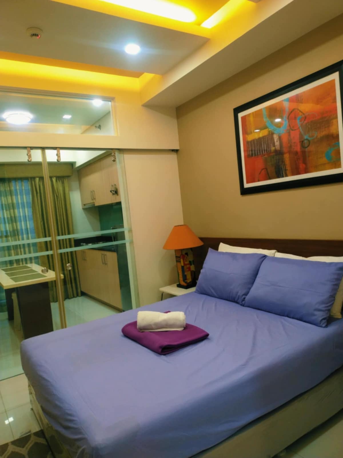 A cozy bedroom features a spacious bed with soft bedding, topped with neatly arranged towels. A bright lamp and framed artwork add color to the neutral walls, while large glass sliding doors provide light and a view into the kitchen area.