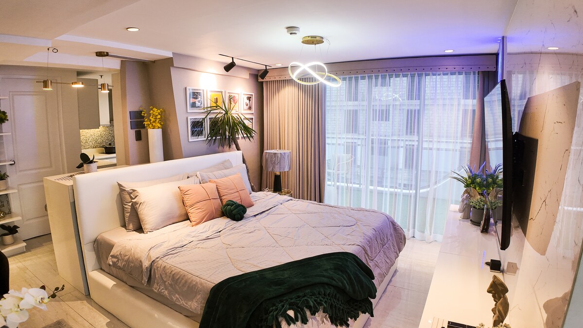 A modern bedroom features a spacious bed with neutral bedding and decorative pillows. Soft lighting is provided by a unique ceiling fixture, while large windows allow natural light to fill the room. An elegant TV stands on the wall opposite the bed, complementing the stylish decor.