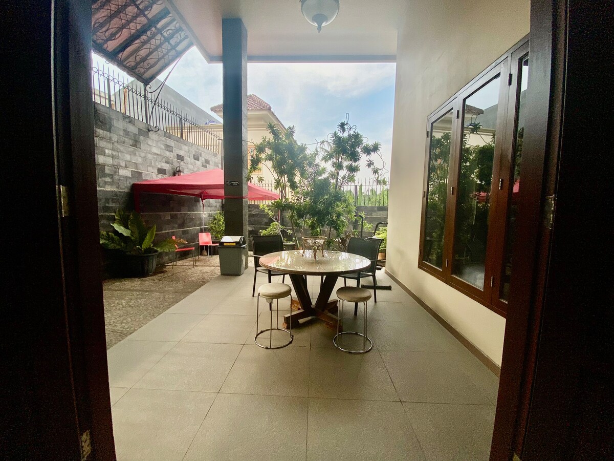 An outdoor dining area is revealed, featuring a round wooden table surrounded by sleek chairs. Lush greenery and potted plants provide a natural backdrop, while a shaded canopy adds protection from the elements. A glimpse of an inviting patio is visible through the open doorway.