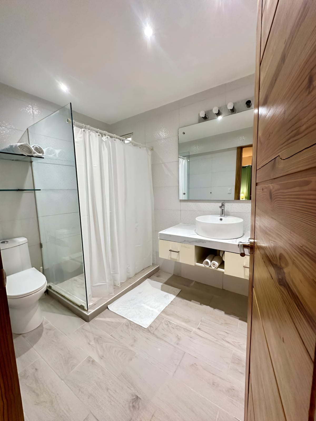 A modern bathroom features a glass-enclosed shower with a white curtain and a sleek vanity with a round sink. Light-colored tiles are complemented by a wooden door and open shelf displaying neatly arranged towels. Soft lighting enhances the clean, functional design.