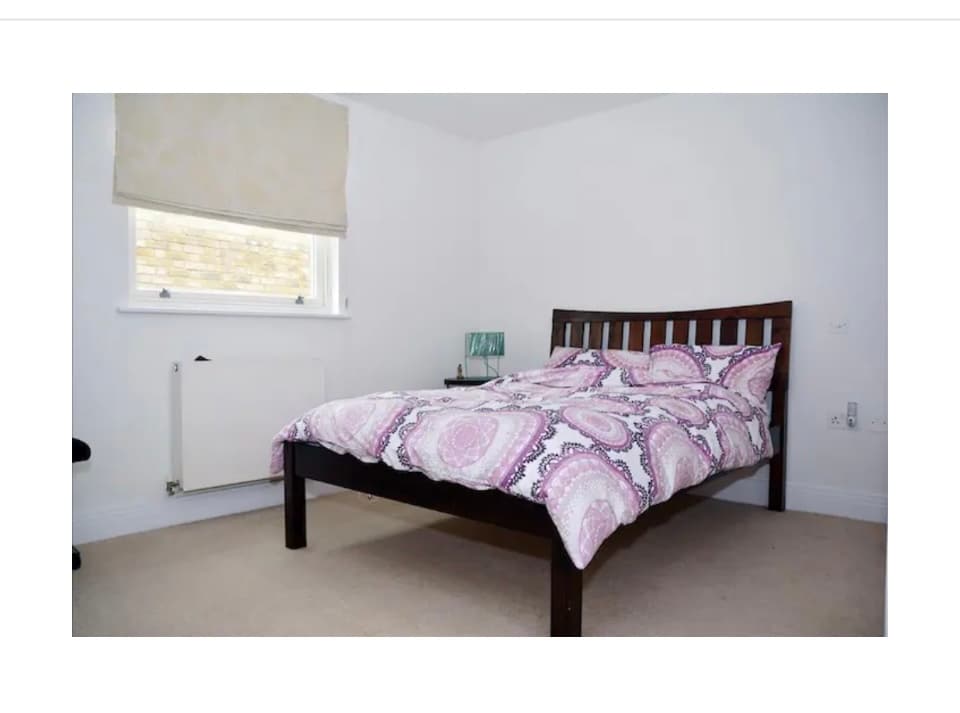 A welcoming bedroom is featured with a spacious bed adorned in a patterned duvet. Natural light enters through the window, which is covered by a light shade. A radiator is positioned below the window, complementing the neutral walls and carpet.
