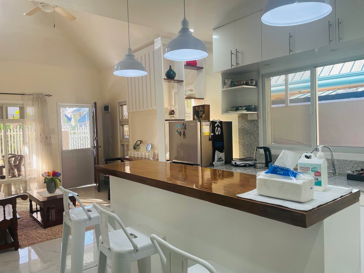 A modern kitchen features a wooden countertop with three white bar stools, illuminated by pendant lights. Stainless steel appliances, including a refrigerator and a kettle, are visible. A window offers natural light, and a cozy dining area is in view, enhancing the inviting space.