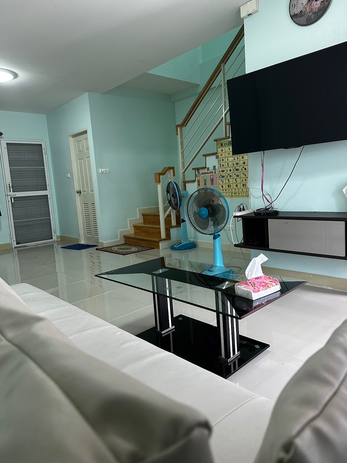 A light-filled living area features a two-tone sofa and a black glass coffee table, accompanied by two ceiling fans. The walls are painted in a soft blue, while a flat-screen television is mounted on the wall. Stairs lead to the upper level, providing additional space.