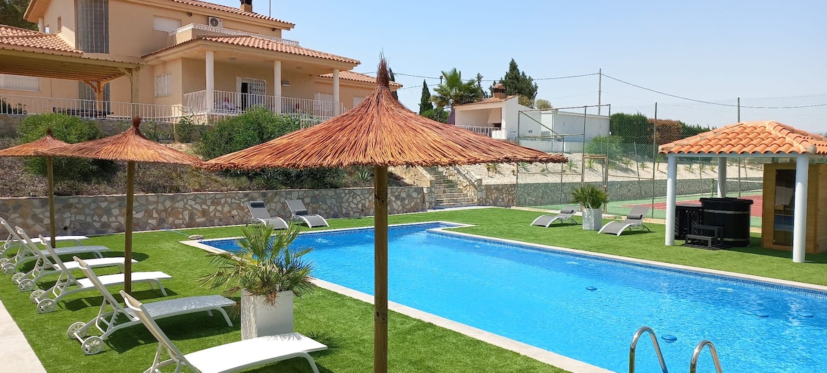 A private swimming pool is set amongst well-maintained grass, accompanied by lounge chairs arranged under thatched umbrellas. The villa's exterior is visible in the background, with a tennis court and landscaped area enhancing the outdoor setting.