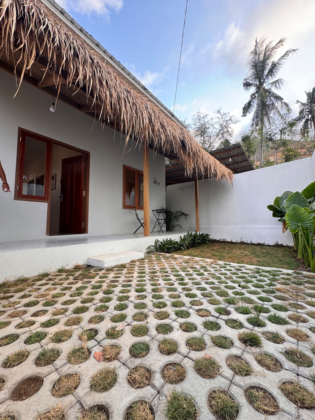 A cozy outdoor space is featured, with a stone pathway leading to a house adorned with thatched roofing. Lush greenery is visible, along with a small table and chairs positioned on a grassy area, creating a welcoming atmosphere for relaxation.