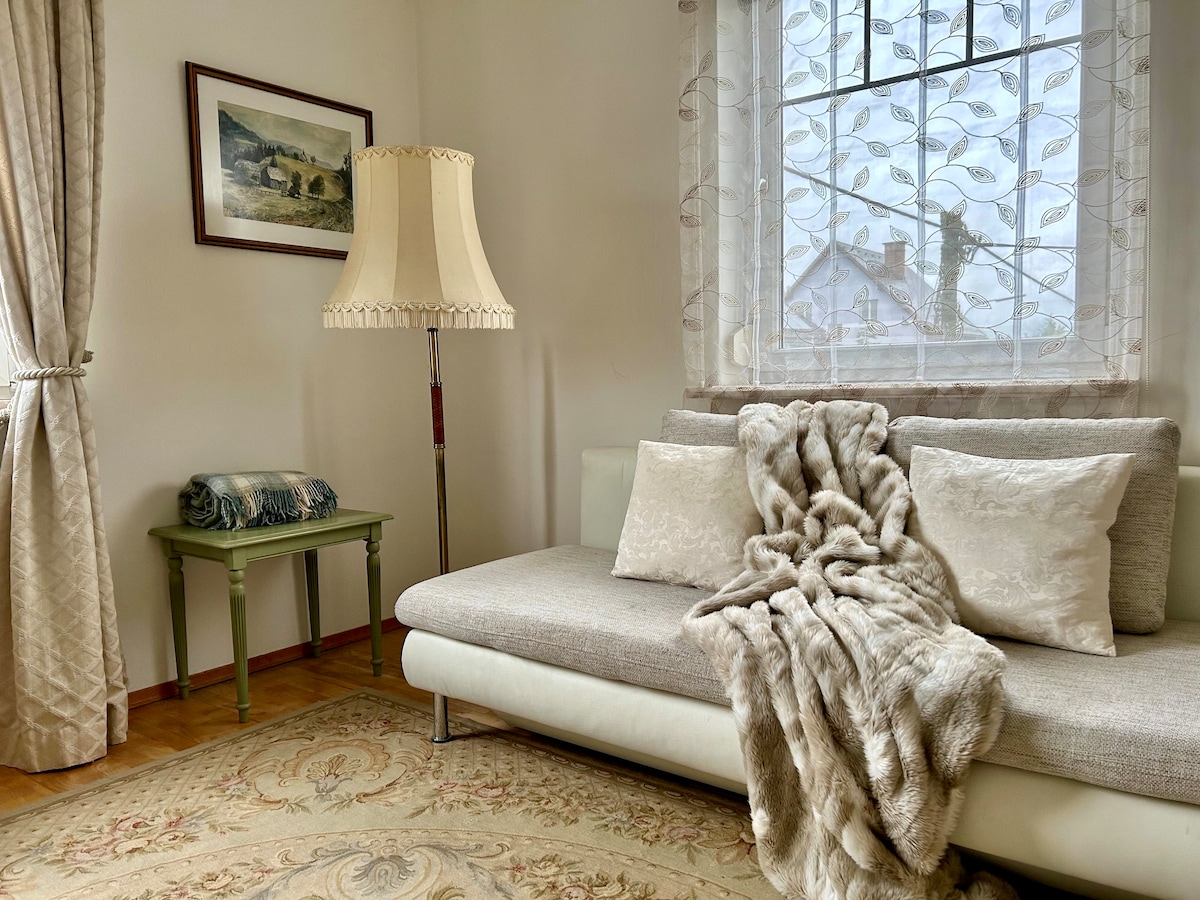 A cozy lounge area features a comfortable sofa draped with a soft throw blanket. A small green table holds a decorative item, while a large window, enhanced by sheer curtains, allows natural light to fill the space. A picturesque artwork adorns the wall.