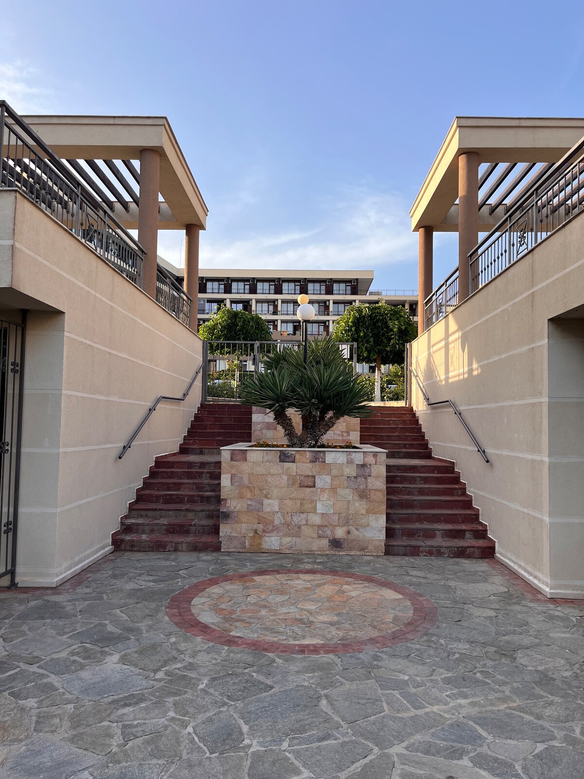 A landscaped entrance features a set of stone steps leading upwards, flanked by decorative planters and greenery. The pathway is made of irregular stones, with a circular stone design at its center. The complex is visible in the background, providing a sense of surrounding community.