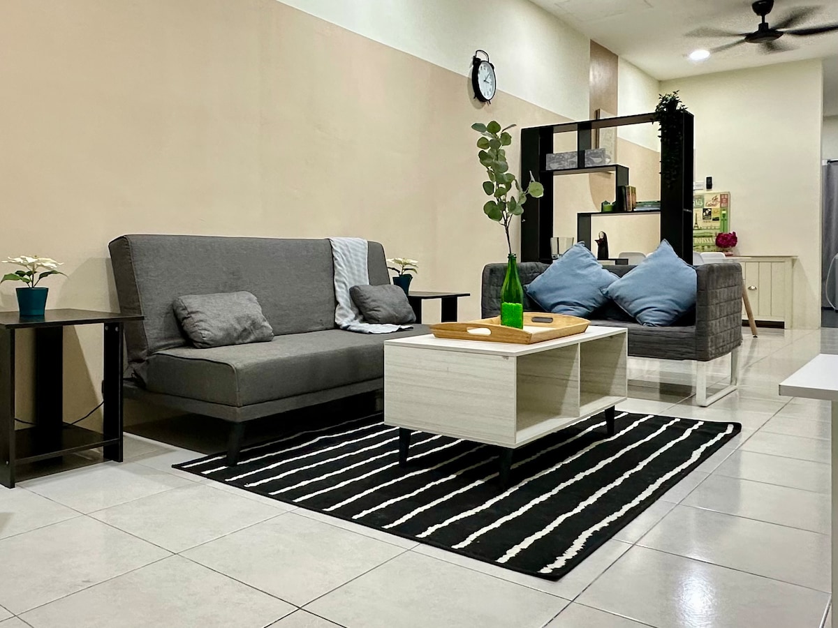 A contemporary living area features a gray couch and two blue accent pillows positioned around a white coffee table. A striped rug anchors the space, while potted plants add touches of greenery. Bright lighting and a neutral wall color enhance the open feel.