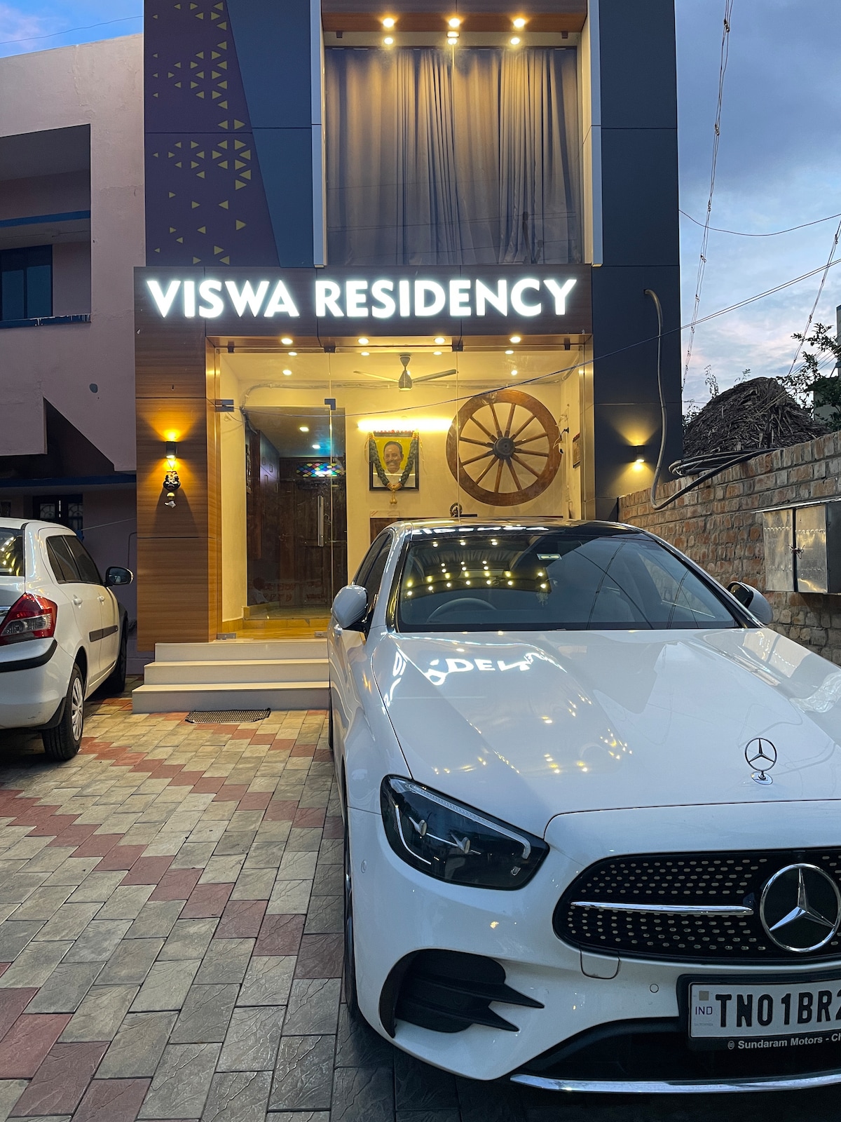 The entrance to Viswa Residency is showcased, featuring a modern facade with illuminated signage. A stylish white car is parked in front on a patterned driveway. Warm lighting highlights the inviting entrance and decorative elements.