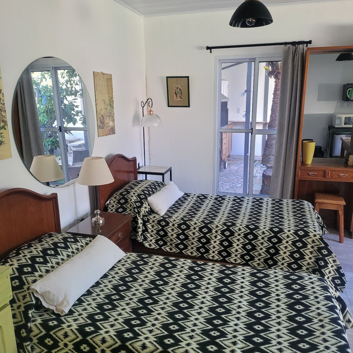 A cozy sleeping area features two beds with patterned bedspreads, accompanied by bedside lamps. A round mirror reflects the natural light entering through the window, which offers a view of greenery. A wooden dresser and a small table contribute to the welcoming environment.