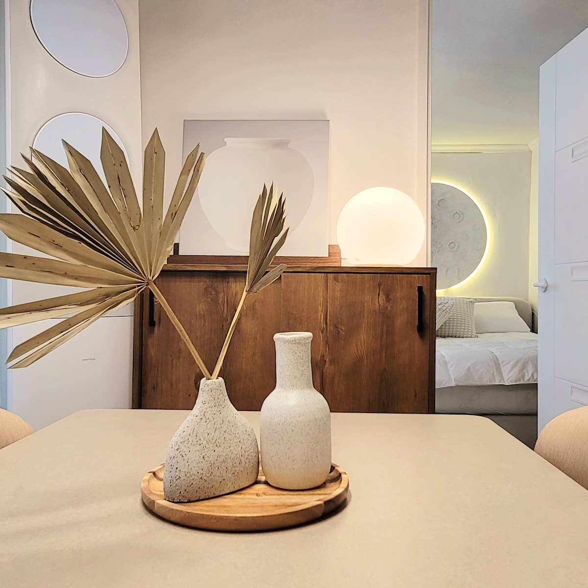 A simple tabletop is adorned with two textured vases and a wooden tray. In the background, a softly lit bedroom is visible, showcasing a round wall lamp and a bed, subtly enhancing the serene ambiance of the space.