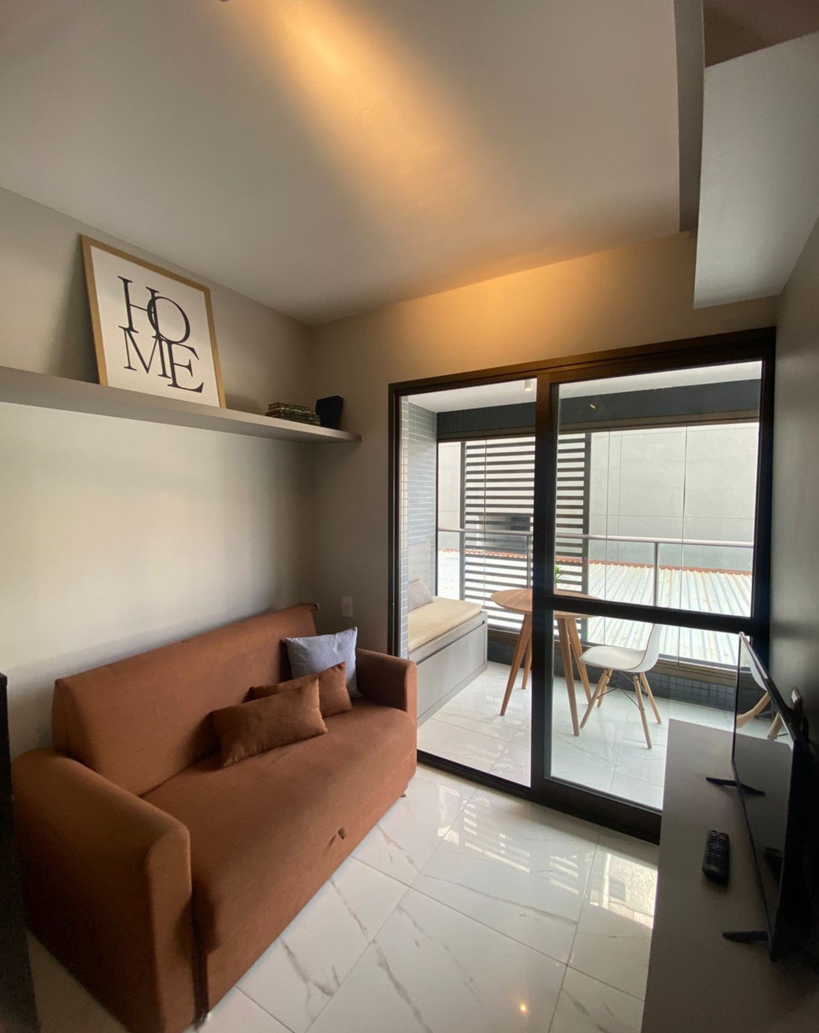 A cozy seating area features a brown sofa adorned with decorative cushions. A large glass door opens to a private balcony space with outdoor seating. Light-colored walls and a shelf with decor create a serene atmosphere, enhanced by natural light filtering in.