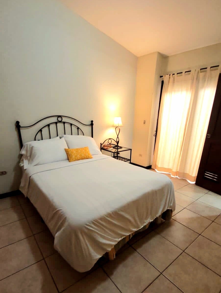 A serene sleeping area is presented with a spacious bed dressed in white linens. A small bedside table holds a lamp next to the bed. Natural light enters through a window with light curtains, creating a calm ambiance.